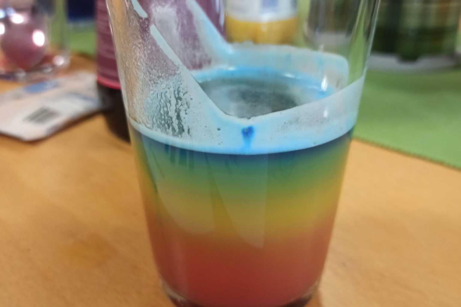 Nahaufnahme des fertigen Rainbow-Cocktails, das Getränkt ist unten rot, darüber orange und gelb und wird über grün zu blau an der Oberfläche