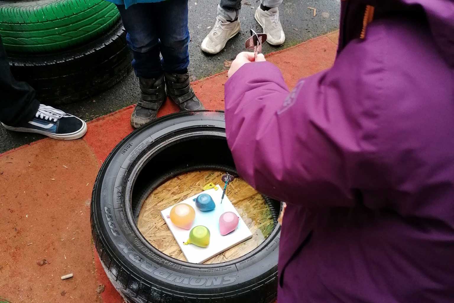Ein Kind versucht mit einem Dart-Pfeil am einem Band Luftballons auf dem Boden innerhalb eines Reifens zu treffen.