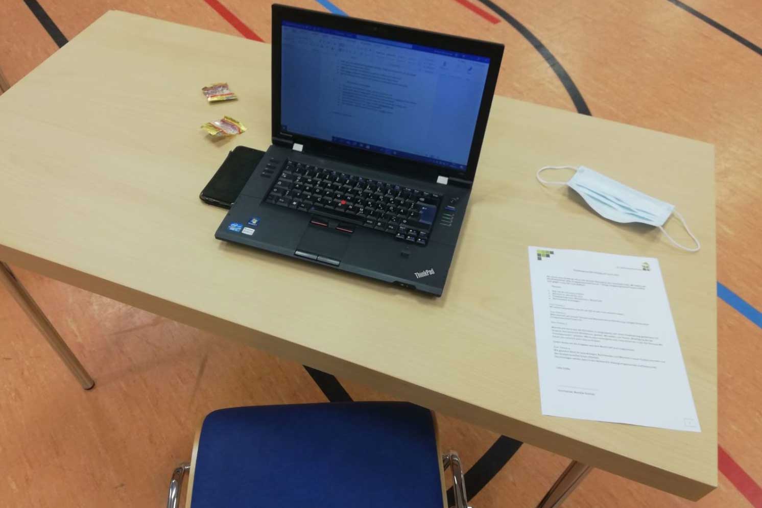 Ein Laptop steh auf einem Tisch, daneben ein Blatt mit der Tagesordnung und eine OP-Maske