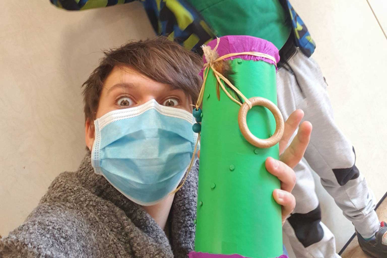 Ein Kind zeigt seinen selbsgemachten Regenmacher, ein Rohr in grün mit lila Deko