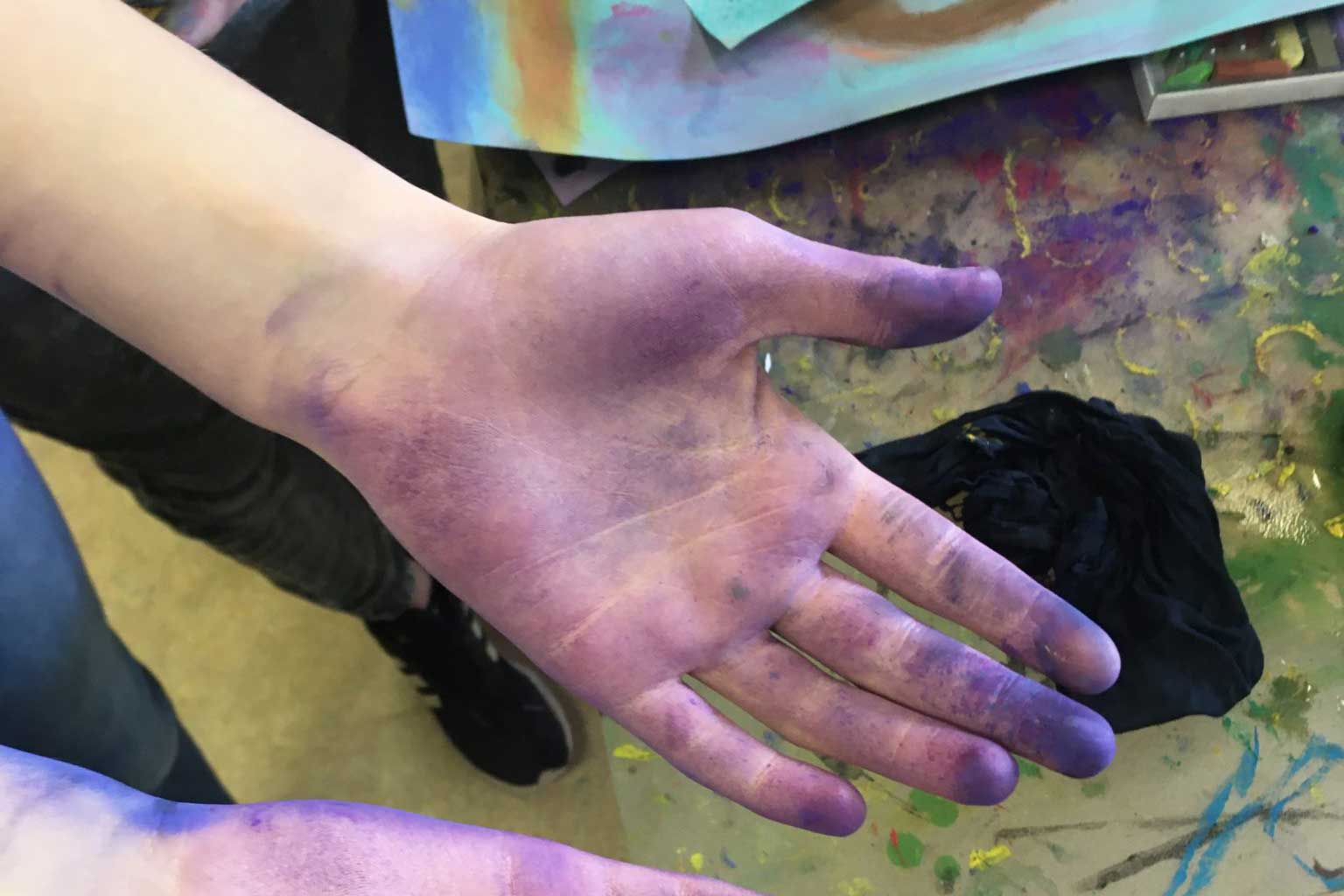 Eine Kinderhand mit lila Farbe, im Hintergrund Bilder und Farbkleckse